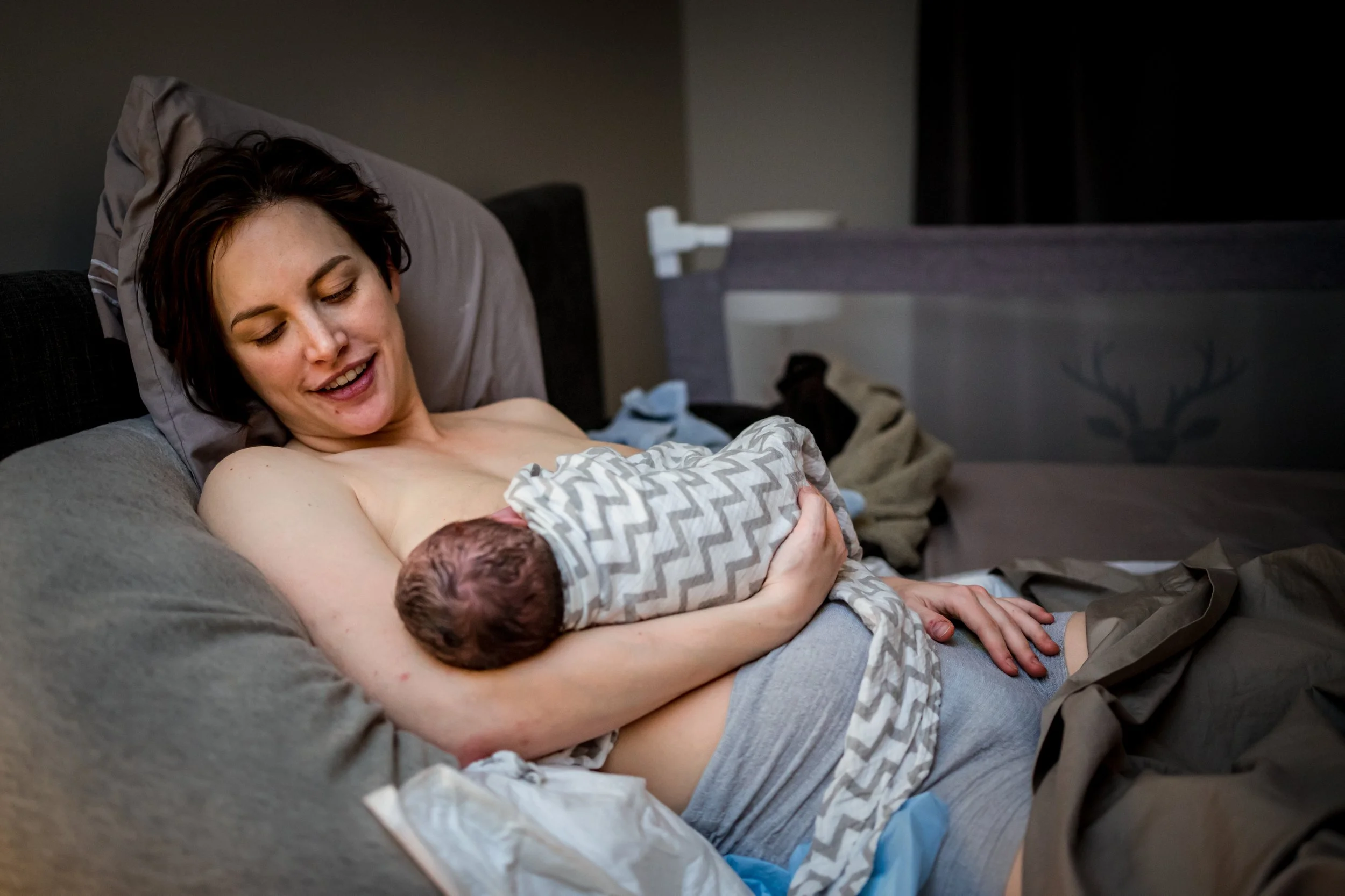 A tired postpartum mother sitting upright in bed on day three after birth, gently breastfeeding a newborn in soft morning light, realistic photo