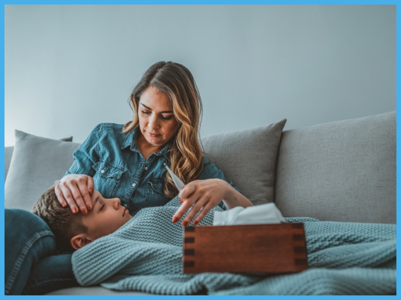 A tired preschool-aged child with flushed cheeks resting under a light blanket on a living room couch while a parent checks their forehead, realistic home photography style
