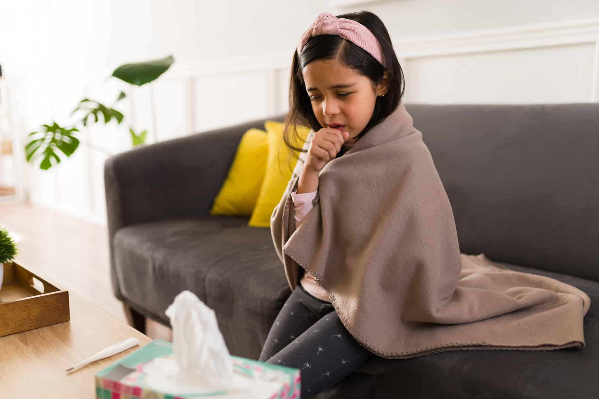 A tired toddler sitting on a living room couch with a parent nearby, coughing into their elbow in natural window light, candid family photograph