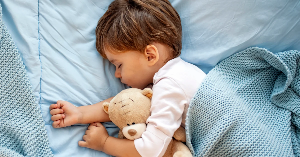 A toddler asleep on a small bed with a light blanket in a softly lit bedroom during daytime, candid lifestyle photograph