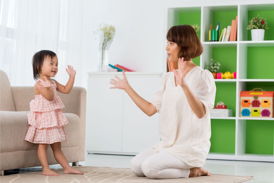 A toddler mid-freeze during a dance game in a cozy living room while a parent watches nearby, candid photo style