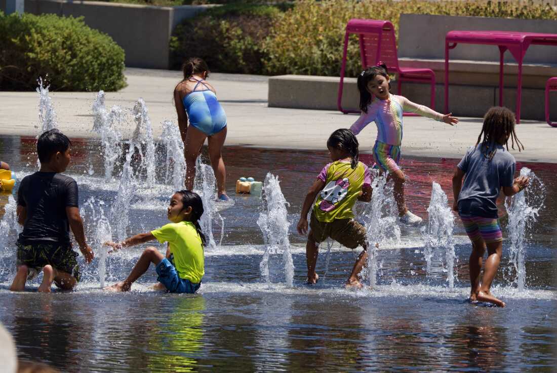 A toddler playing with water toys in a shaded backyard on a sunny day, wearing a sun hat and light clothing, realistic photo