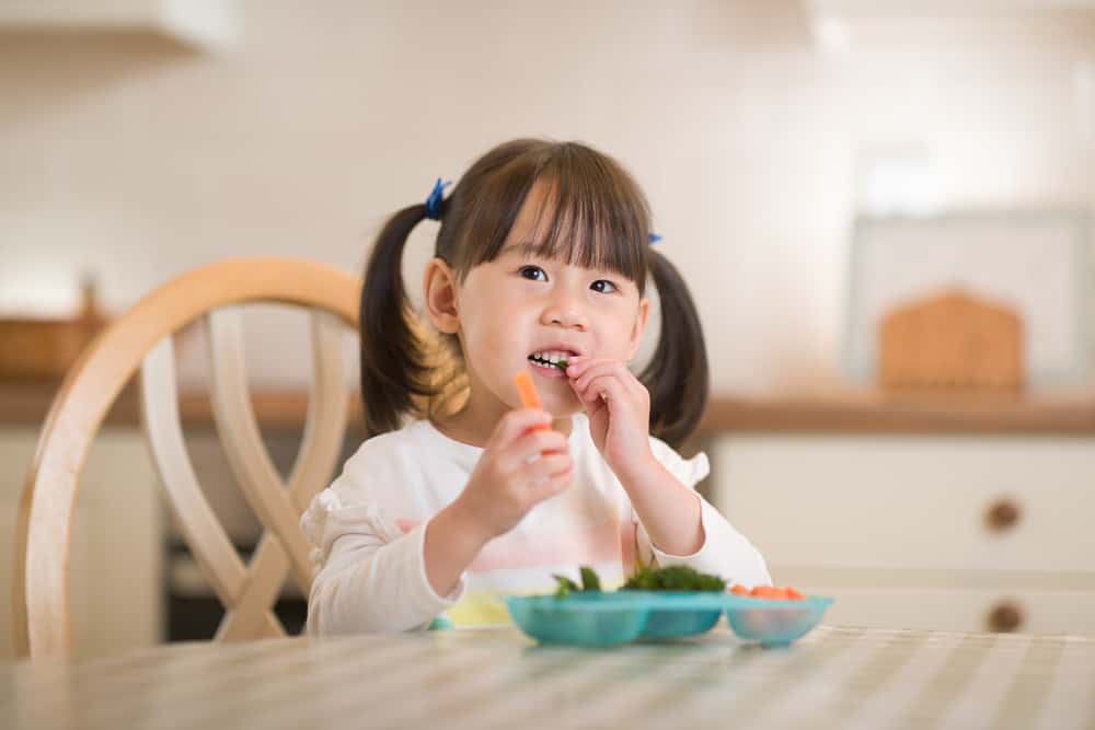 A toddler sitting at a kitchen table eating a snack while a parent watches nearby with a glass of milk, warm natural light photo