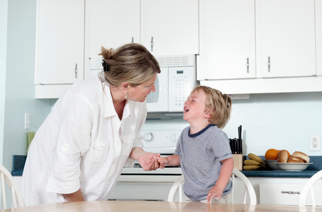 A toddler sitting at a kitchen table looking up while talking to a parent who is leaning in to listen, natural indoor family photo