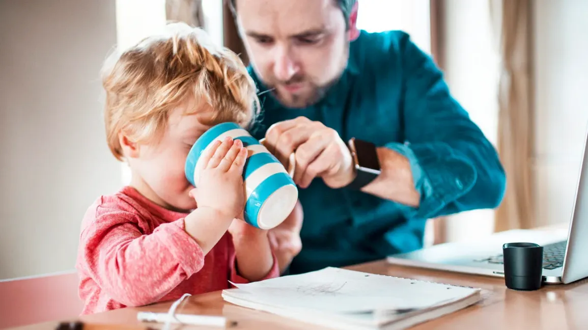 A toddler sitting at a kitchen table sipping a warm caffeine-free drink from a spill-proof cup while a parent watches closely, cozy indoor photo