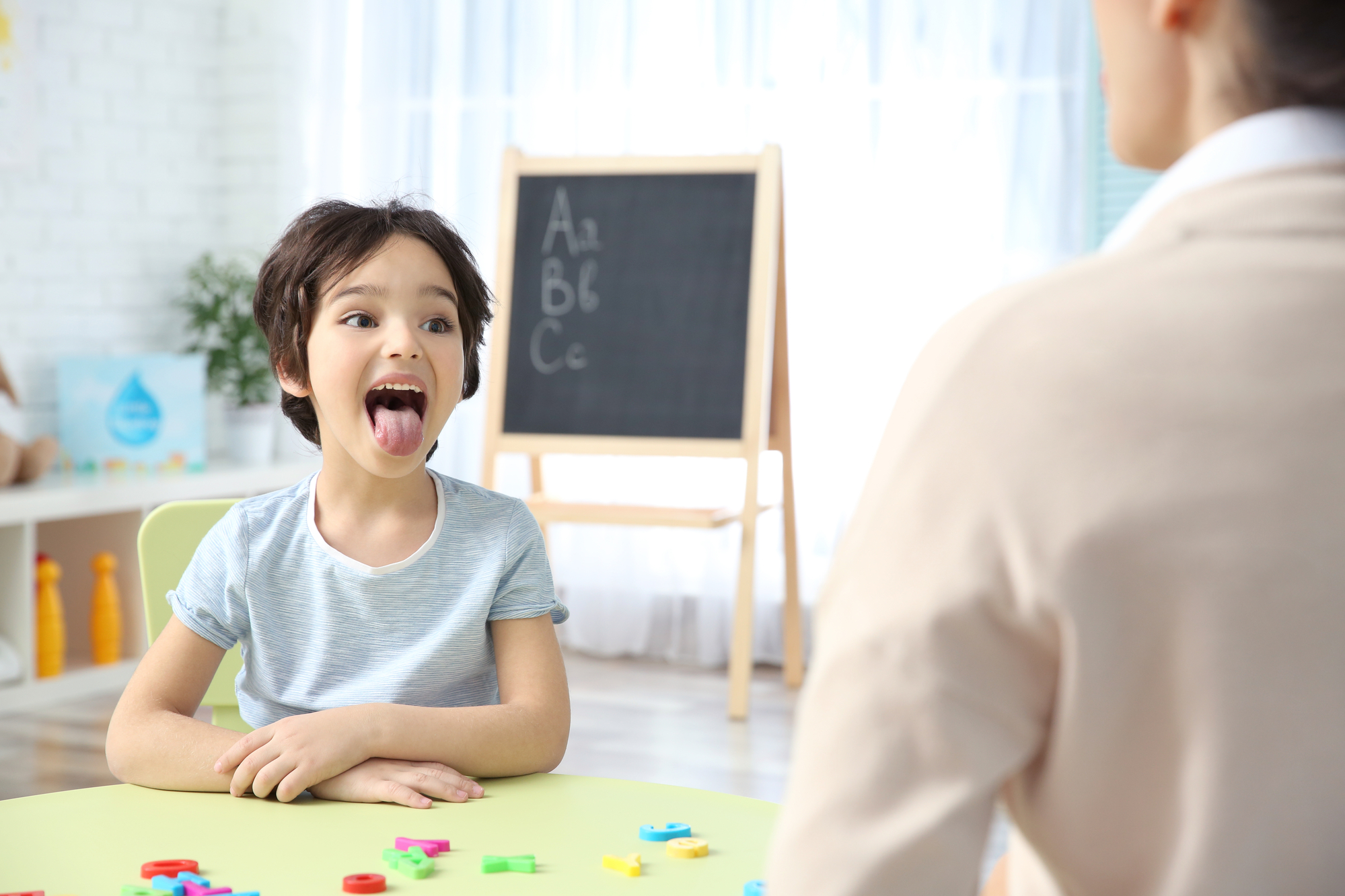 A toddler sitting at a small table in a pediatric speech therapy room while a speech-language pathologist models simple sounds with picture cards, natural indoor light