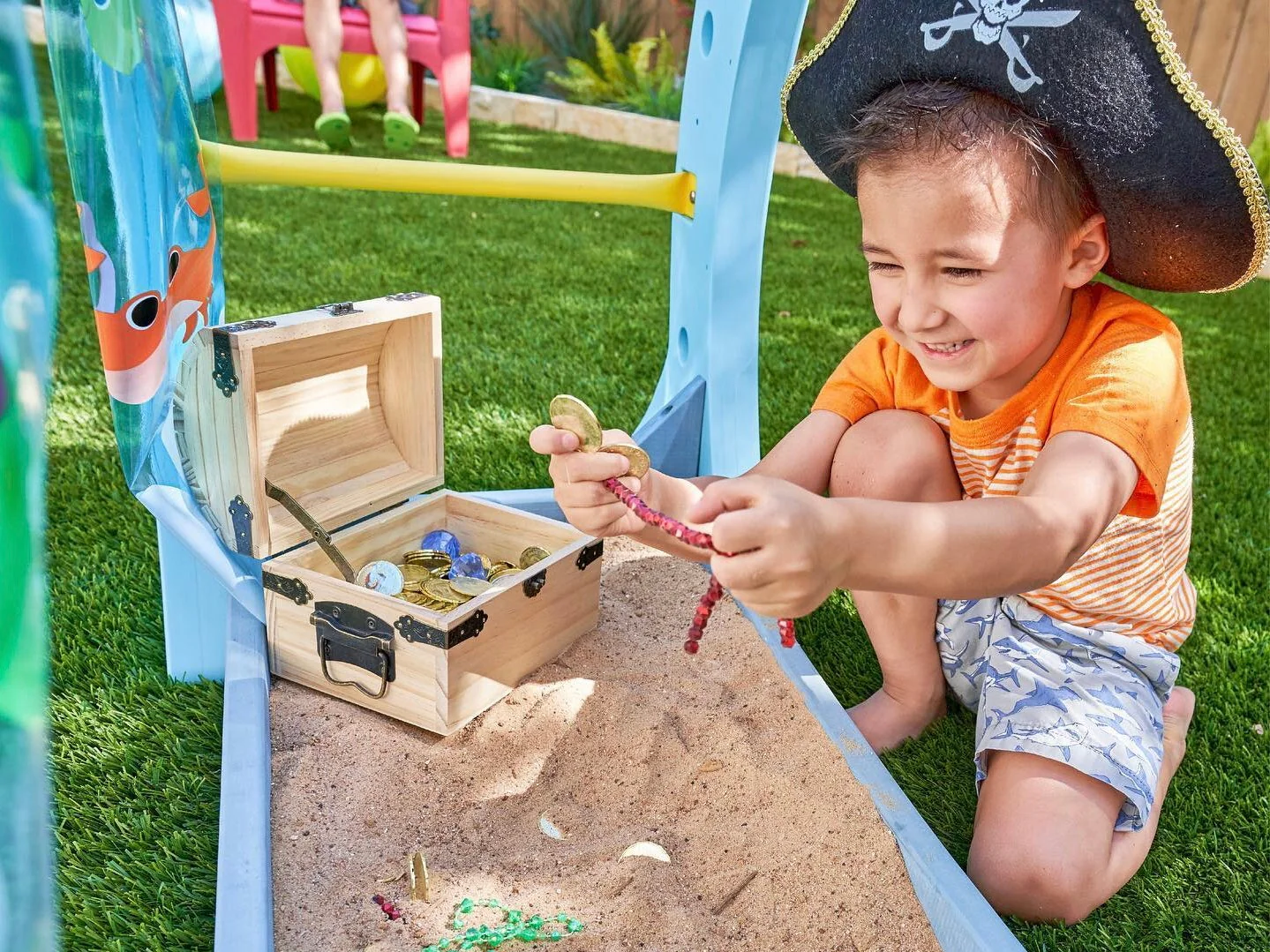 A toddler sitting in a daycare sandbox outdoors with a caregiver in the background, natural candid photo style
