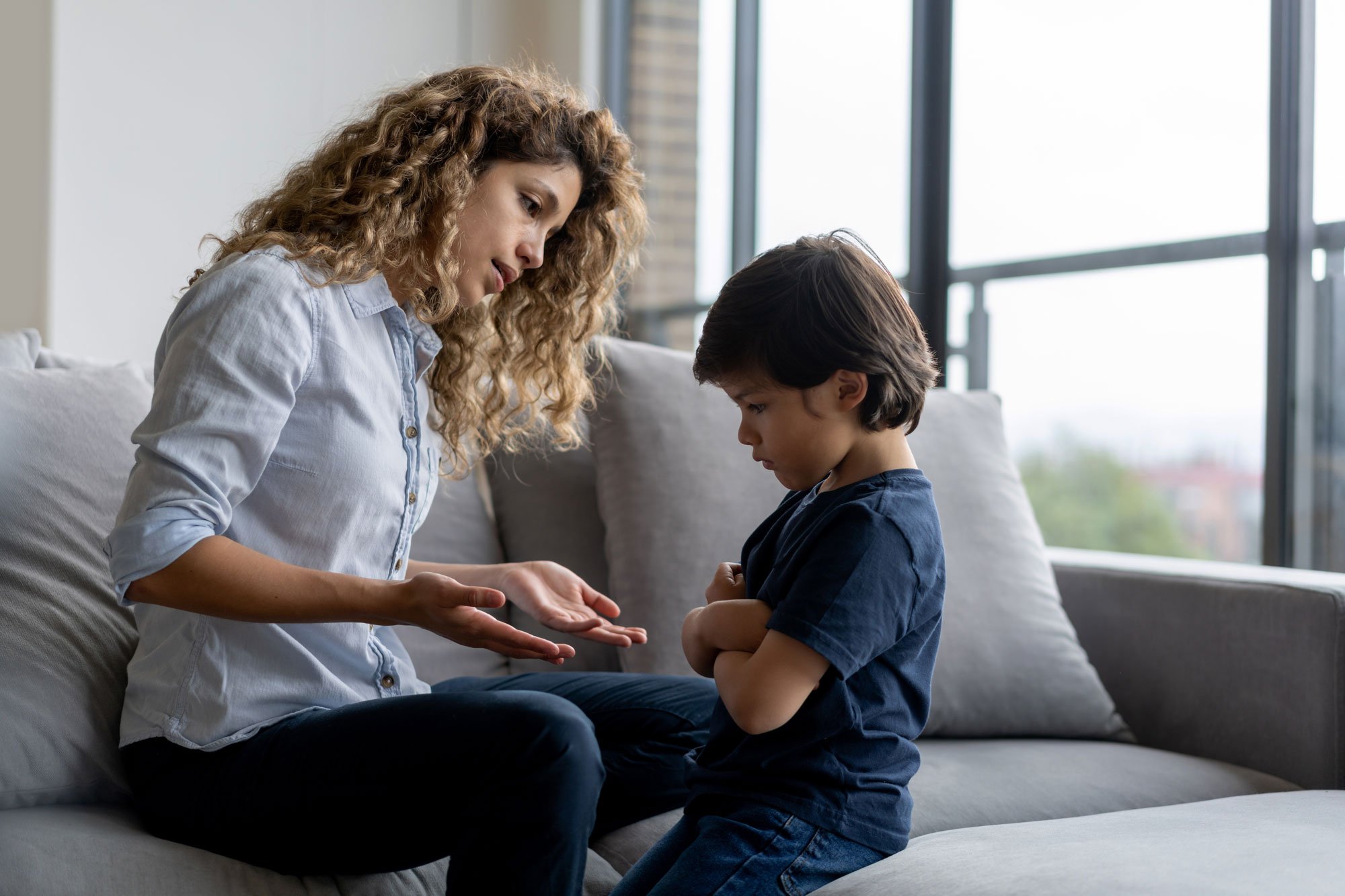 A toddler sitting on a living room floor mid-tantrum while a parent kneels nearby with a calm, supportive posture, everyday home setting, real photograph style