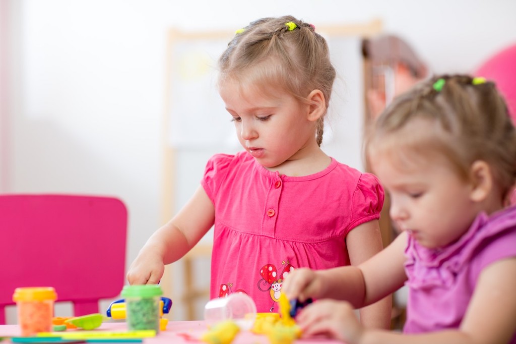 A toddler sitting on a living room rug holding a small plastic bead near their face while a parent’s hand reaches in to gently stop them, real-life photo