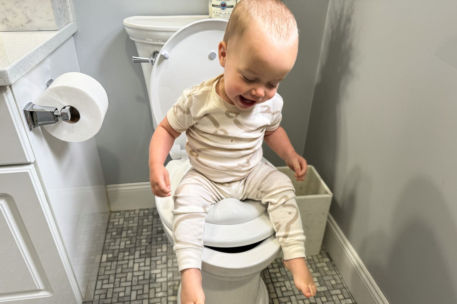 A toddler sitting on a small potty in a bathroom with feet supported on a step stool, natural light, real photograph