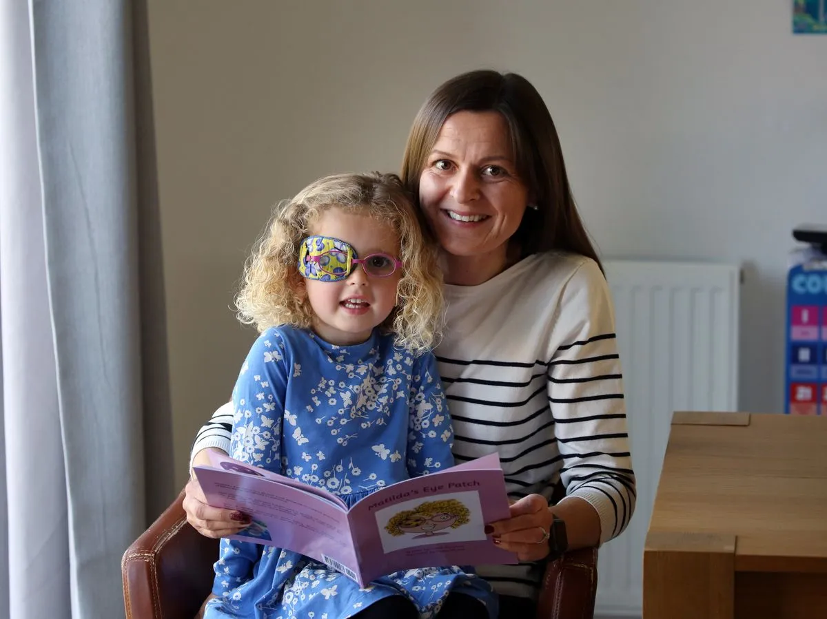 A toddler wearing a soft adhesive eye patch while sitting with a parent reading a picture book on a couch, warm home photography
