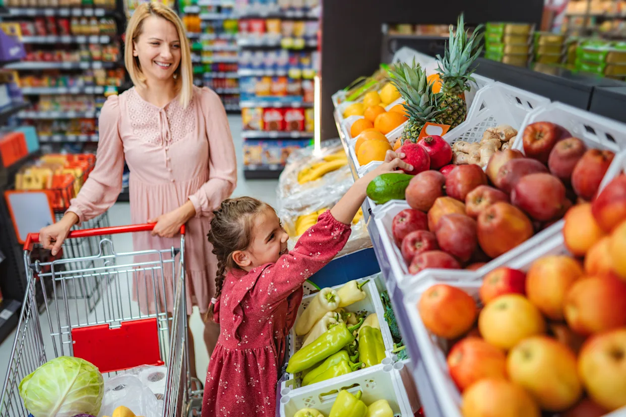 A young child covering their ears in a bright, busy grocery store aisle while a parent stands nearby with a shopping cart, realistic candid photograph