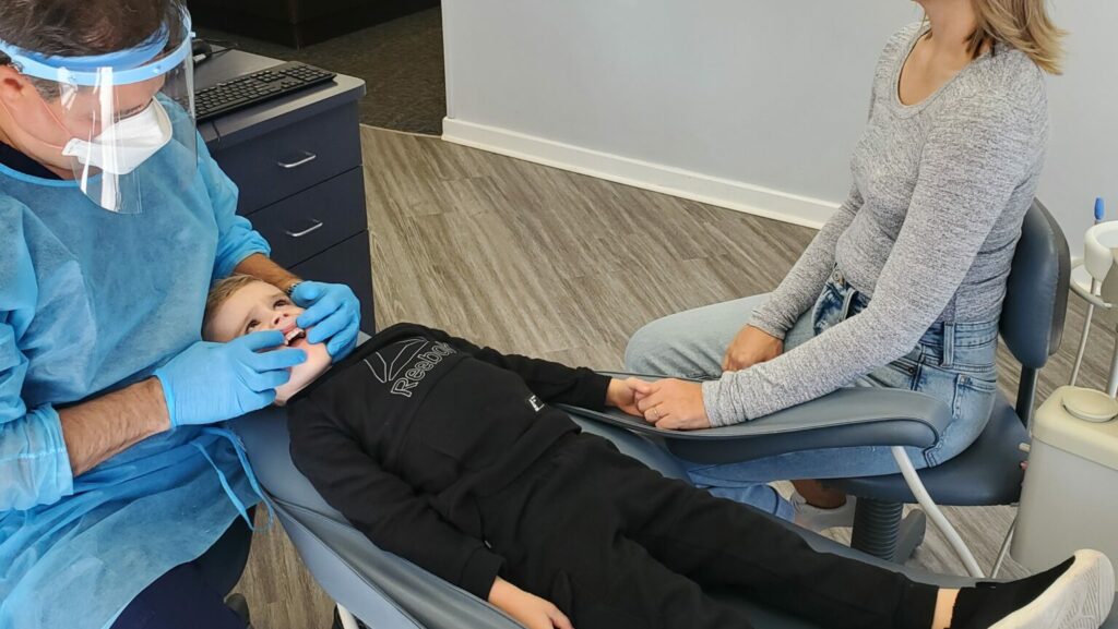 A young child sitting in a dental chair holding a parent’s hand while a dentist prepares to examine their mouth, bright clinical photo