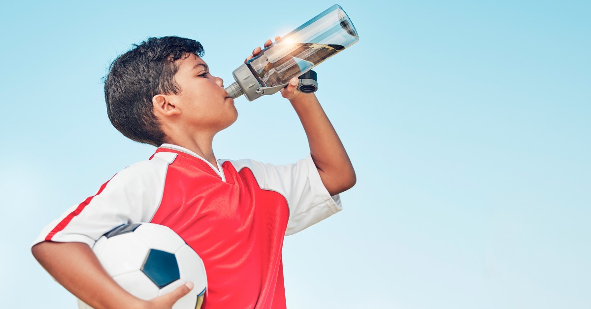 An elementary-aged child in athletic clothes taking a drink from a reusable water bottle on the sidelines of a sports field in late afternoon light, candid photo