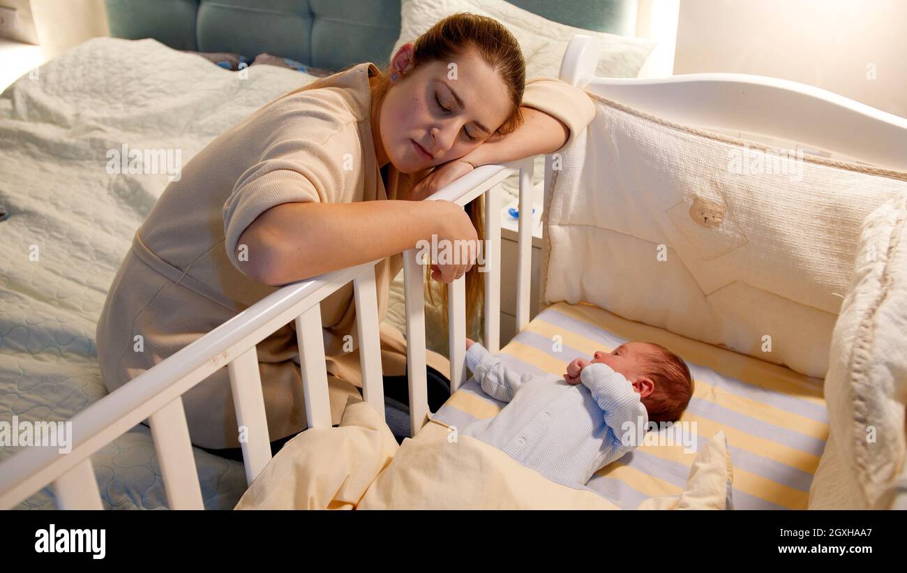 An exhausted parent sitting on the edge of a bed holding a six-month-old baby in a dark bedroom lit by a small nightlight, realistic documentary photo